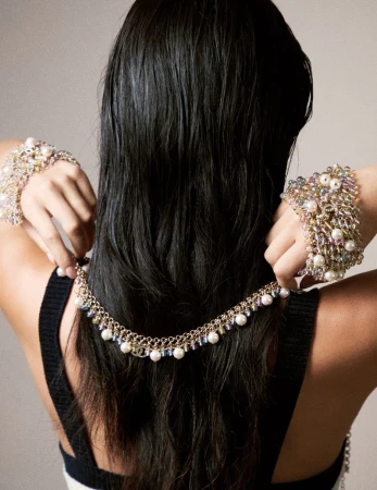 A model displays a gold necklace and pearl bracelets, with her black hair cascading down her back, set against a plain background.