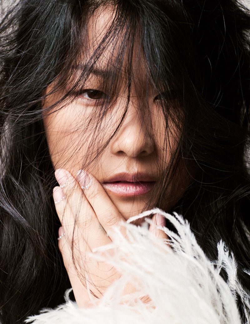 Portrait of an adult woman with dark hair partially covering her face, adorned with white feathers around her hand and lower face.