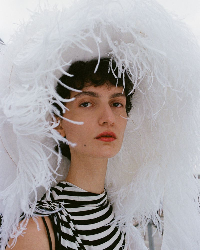 Fashion portrait of a woman wearing a black and white striped top and a large white feathered hat, set against a bright, airy background.