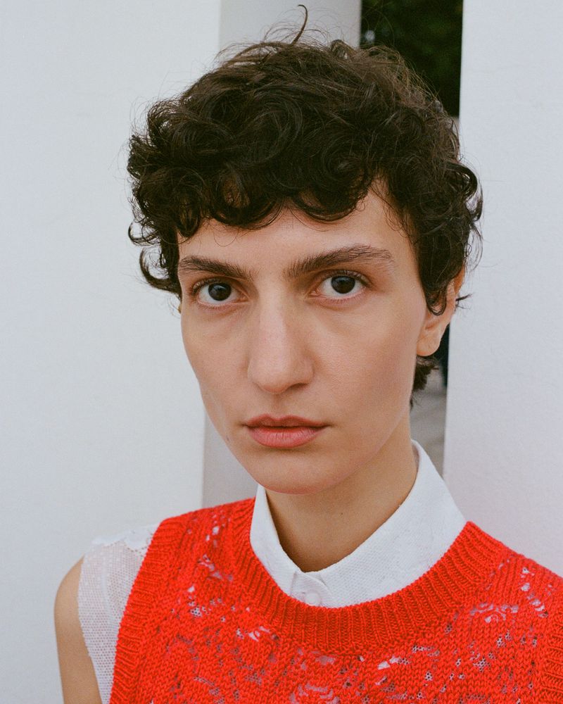 Close-up studio portrait of a woman with curly dark hair wearing a bright orange knitted top over a white collared shirt.