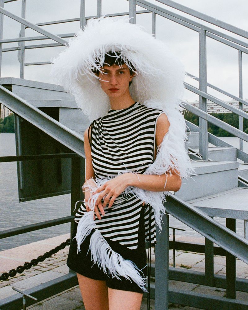 Young woman in striped top and shorts posing with a large white feathered hat outdoors with an urban waterfront and bridge backdrop.