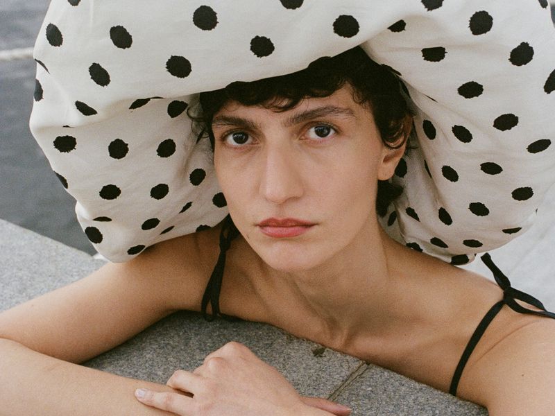 Close-up of a woman wearing a large white hat with black polka dots, looking directly at the camera in natural lighting.