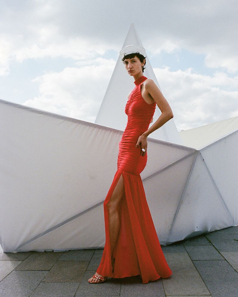 A slender woman in a red gown poses outdoors on a paved surface with angular white architectural structures in the background under a bright sky.