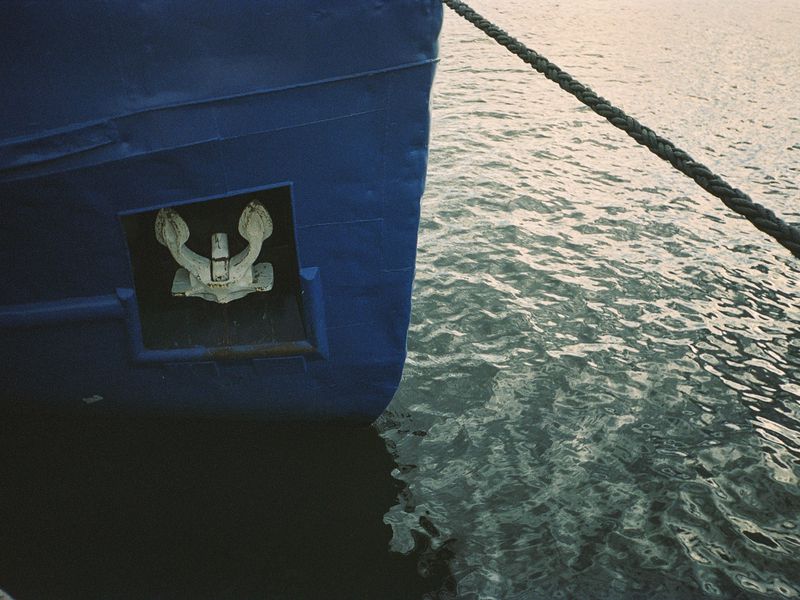 View of the blue bow of a boat with a white anchor, a gray rope, and rippling water in the background, shot from a low angle.