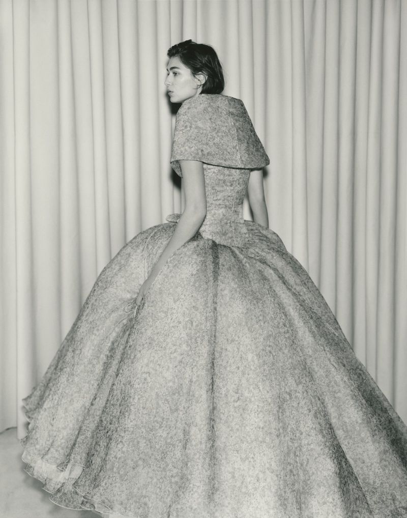 A woman in an ornate ballgown poses in front of a curtain backdrop, showcasing vintage-inspired fashion and design, capturing a timeless aesthetic.