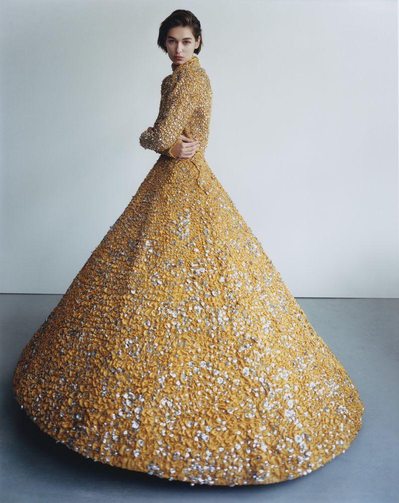 Woman poses in a long-sleeved, gold, textured formal dress with silver embellishments against a plain, light-grey studio backdrop and floor.