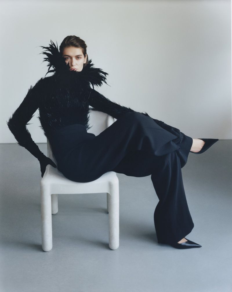 A fashion shot of a woman in a black feather top and wide-legged pants, sitting on a white chair in a photo studio, gazing directly at the camera.