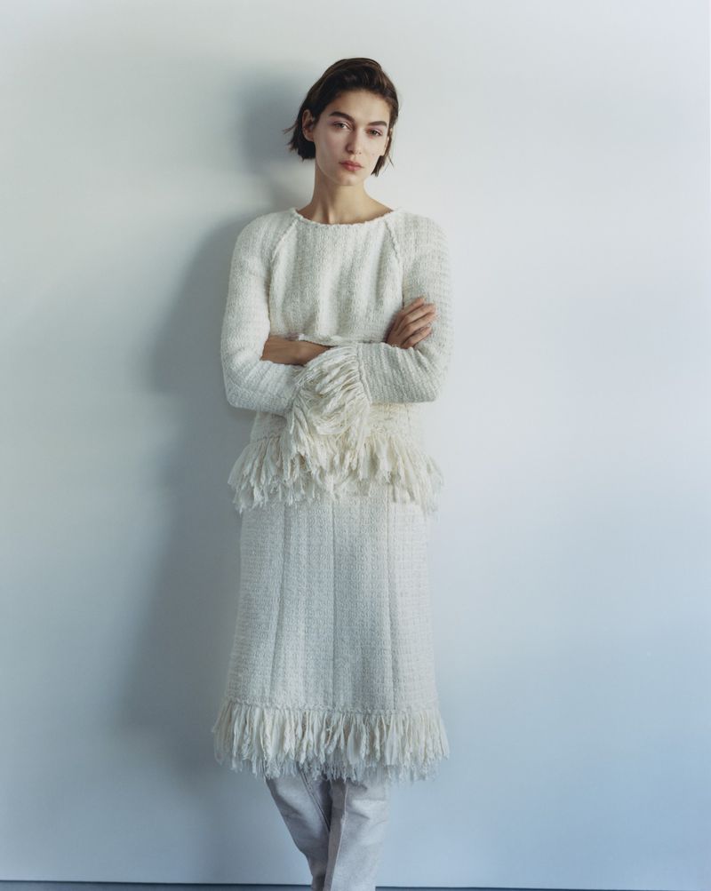 Model Rayssa Medeiros wearing a white textured fringed knit dress and top with arms crossed against a plain light wall.