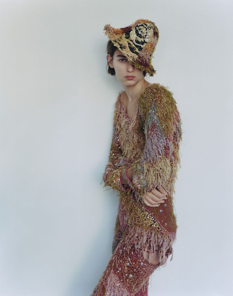 A person poses in a studio wearing a patterned outfit including a hat and long sleeve top and pants with fringe and embellishments.