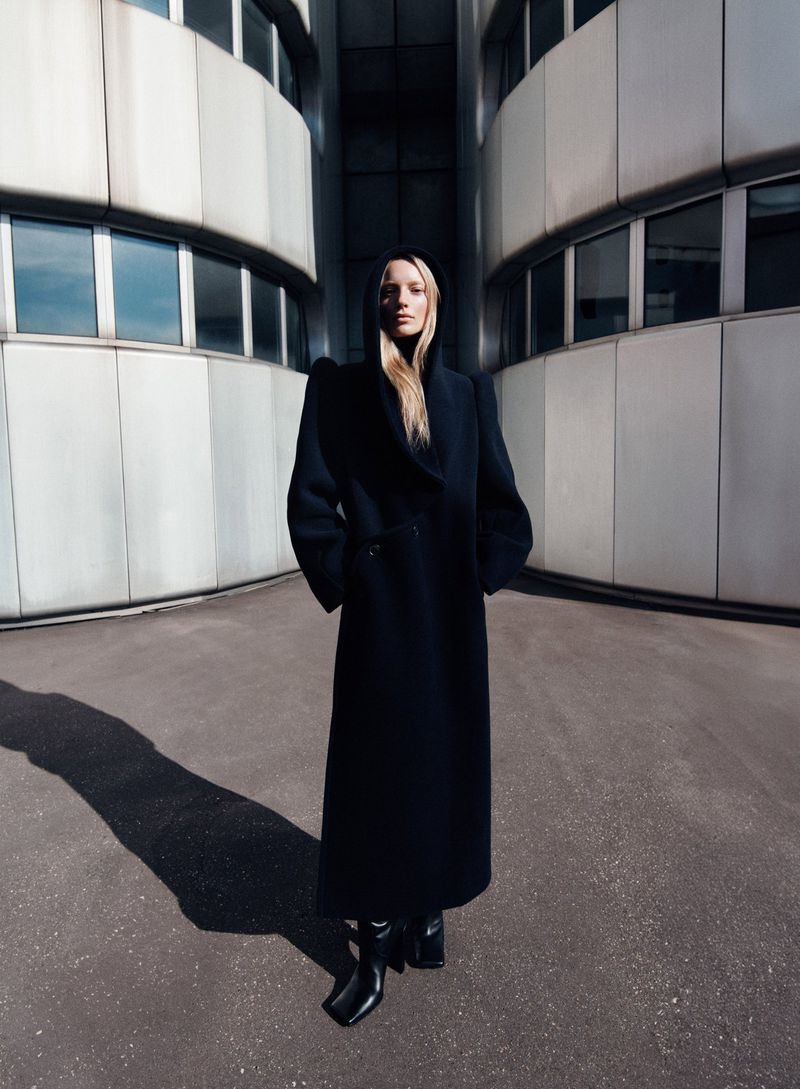 Blonde woman wearing a long black coat with hood, posing outdoors with modern concrete building in background and long shadow on the ground.