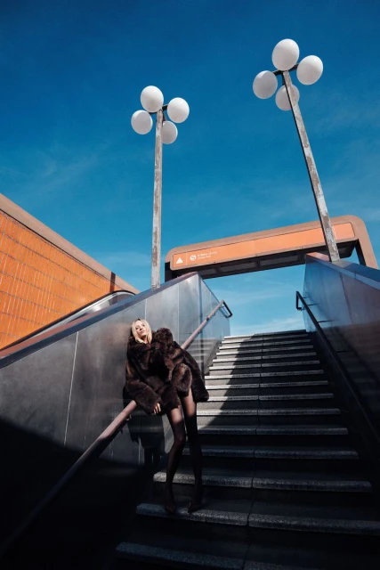 A woman in a brown fur coat sits on staircase railing under streetlights against a blue sky in an urban setting on a sunny day.