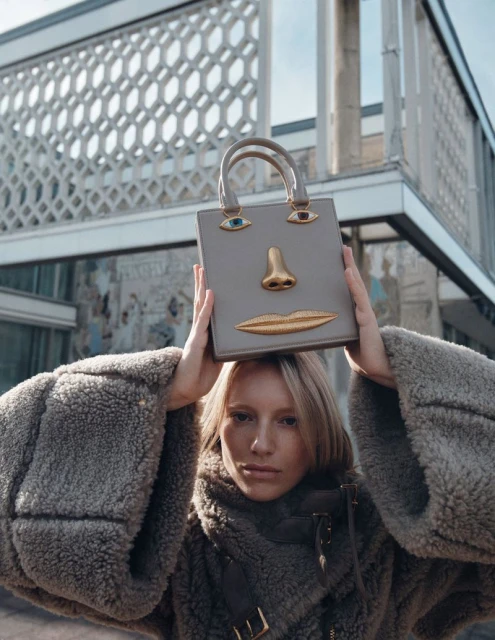 A fair-skinned woman in a furry coat stands outdoors, holding a gray handbag with decorative face-like details, against a building backdrop.