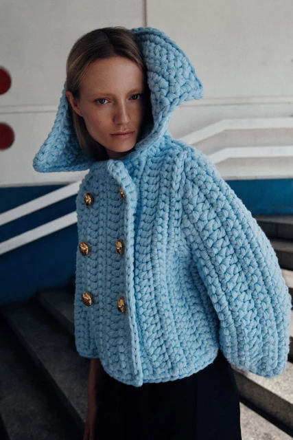 A woman in a blue knitted jacket with gold buttons is posing against a blue and white wall with geometric details indoors.