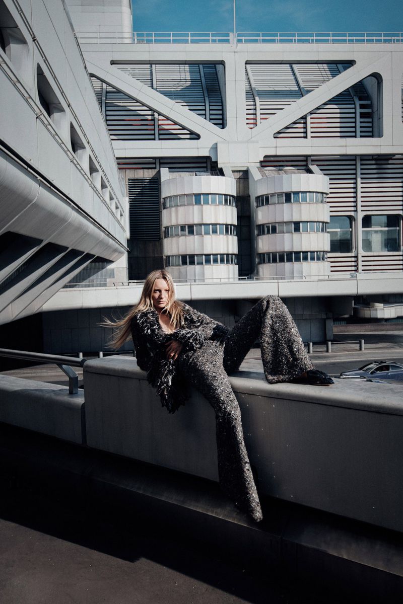 Fashion shot of a blond woman in a sparkling pants suit sitting on a ledge with a large architectural building in the background on a sunny day.