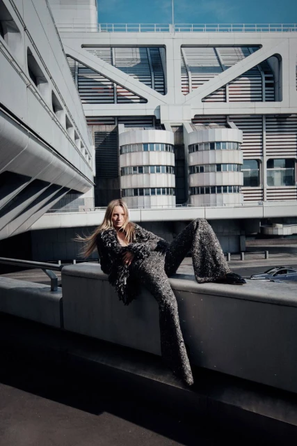 Fashion shot of a blond woman in a sparkling pants suit sitting on a ledge with a large architectural building in the background on a sunny day.