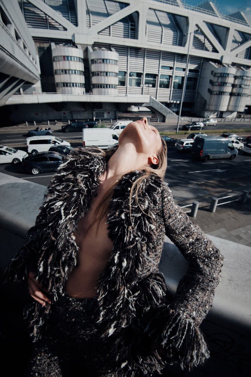 Woman in a textured jacket looks up at a modern building with cars on the road. Fashion shot with neutral, desaturated colors.