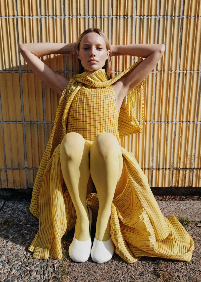 Blonde woman poses with hands behind head, wearing a yellow pleated dress, leggings, and white shoes, sitting against a yellow textured wall.