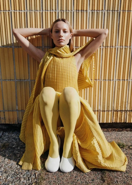 Blonde woman poses with hands behind head, wearing a yellow pleated dress, leggings, and white shoes, sitting against a yellow textured wall.