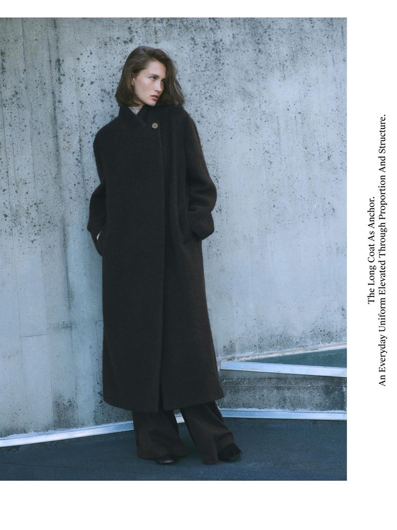 A woman models a dark, oversized wool coat and wide-leg pants, posing against a textured concrete wall in a minimalist fashion shot.