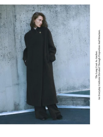 A woman models a dark, oversized wool coat and wide-leg pants, posing against a textured concrete wall in a minimalist fashion shot.