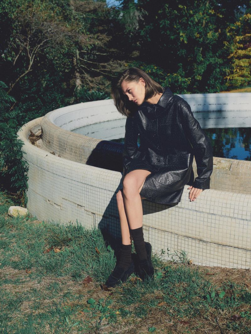 Woman in a black dress sits on the edge of a round, mosaic-tiled fountain in a grassy outdoor setting with a background of trees.