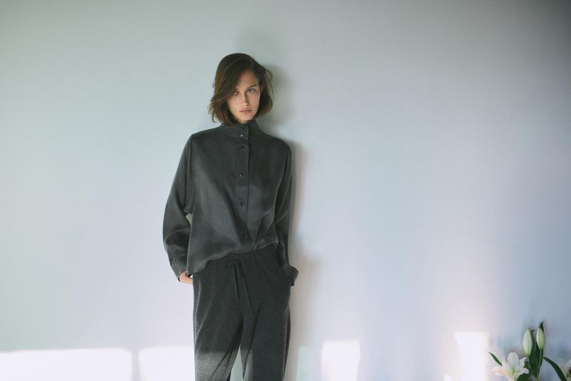 Model Vika Evseeva wearing a charcoal grey high-neck button-up shirt and relaxed trousers against a pale wall.