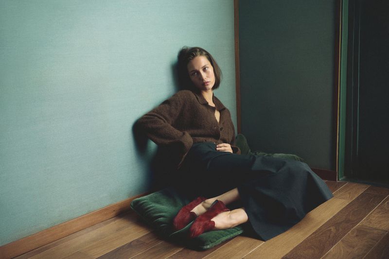 A young woman with dark hair poses indoors wearing a brown sweater, black pants, and red shoes, sitting on a green cushion against a blue wall.
