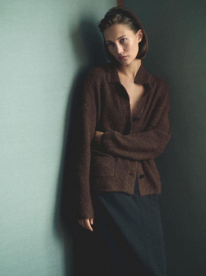 Young woman in a brown cardigan leaning against a green wall, medium shot, soft lighting, fashion editorial style, minimal background.