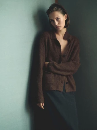 Young woman in a brown cardigan leaning against a green wall, medium shot, soft lighting, fashion editorial style, minimal background.