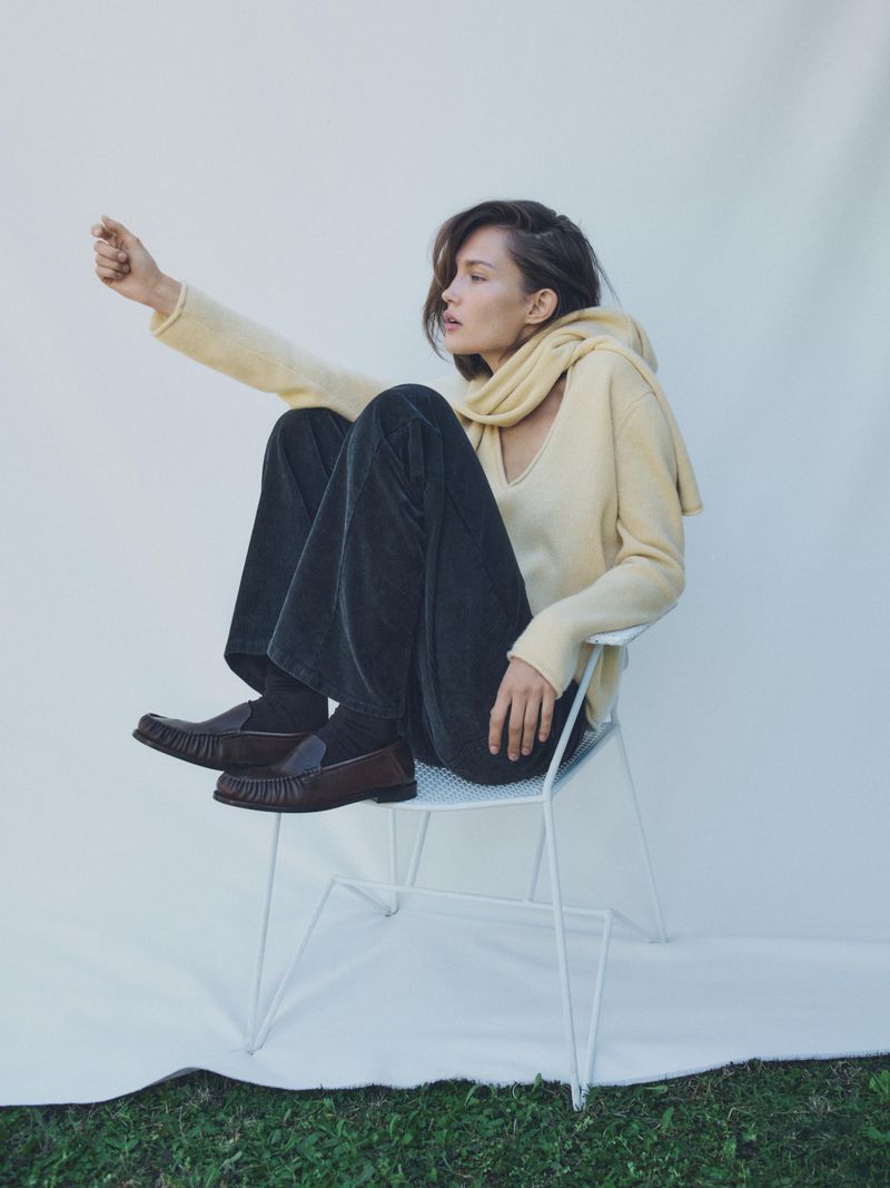 Young woman sits on a white chair in a photo studio, wearing casual sweater, pants, and loafers, gazing out of frame.