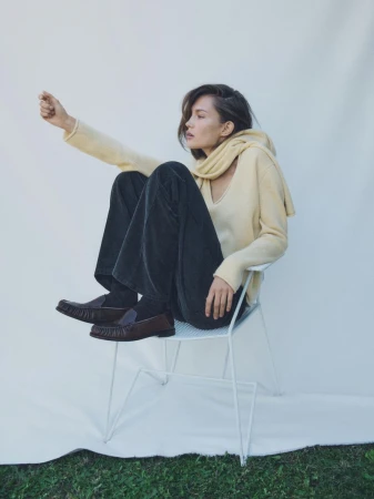 Young woman sits on a white chair in a photo studio, wearing casual sweater, pants, and loafers, gazing out of frame.
