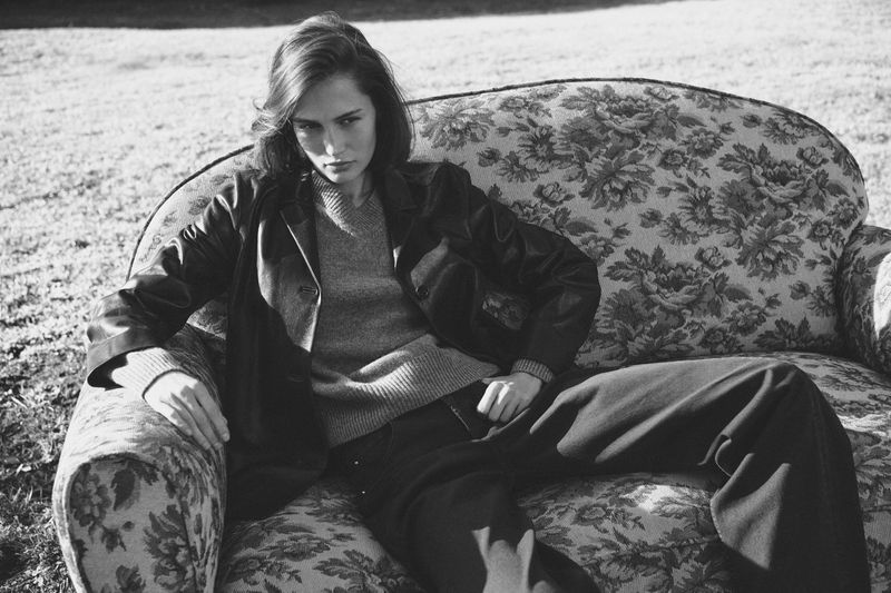 A woman with brown hair lounges on a floral couch in a field. She wears a sweater, blazer, and slacks in this black and white fashion shot.