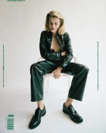 Sylvia Peterson sits on a white chair wearing a black leather jacket and pants with black shoes in a fashion editorial shot.