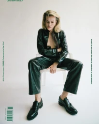 Blonde woman seated on a white chair wearing a black leather jacket and matching pants against a white background