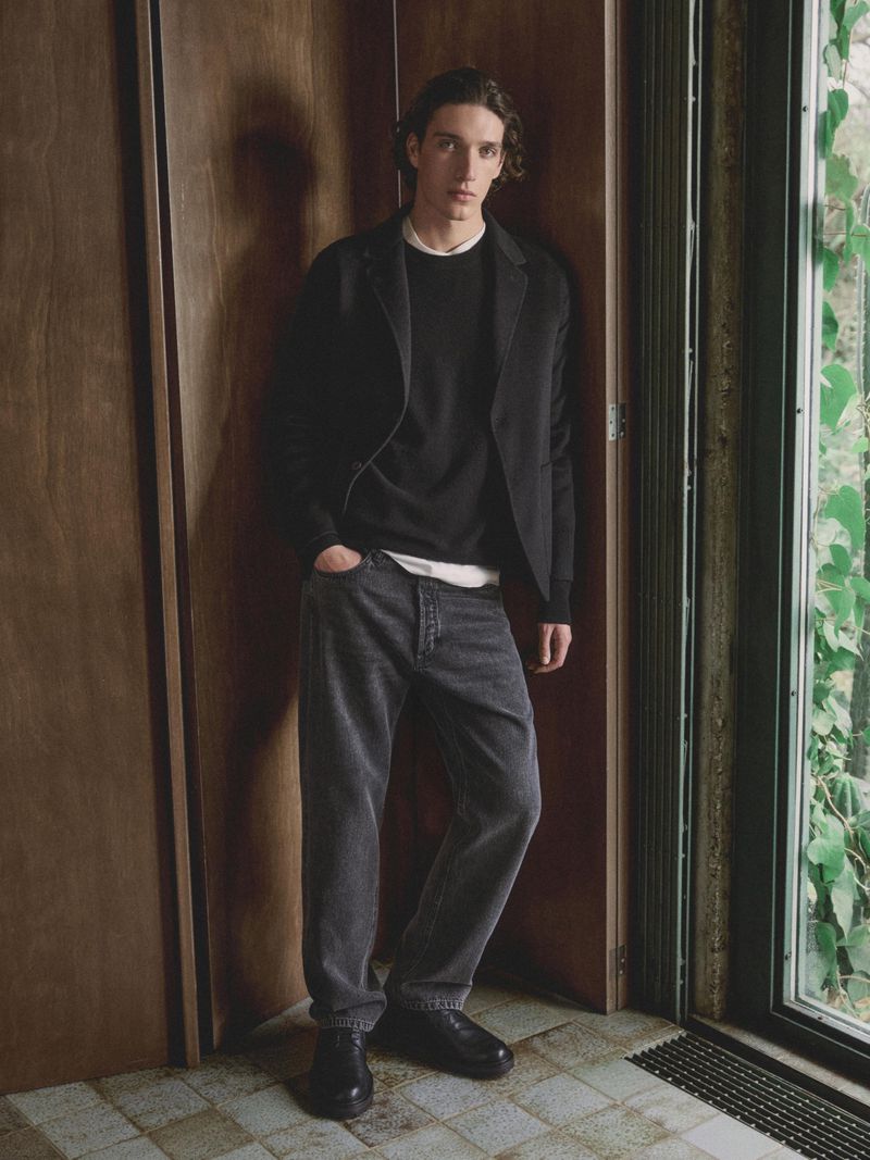 Male model poses indoors wearing a black blazer, white t-shirt, grey jeans, and black leather boots, leaning against a paneled wall.