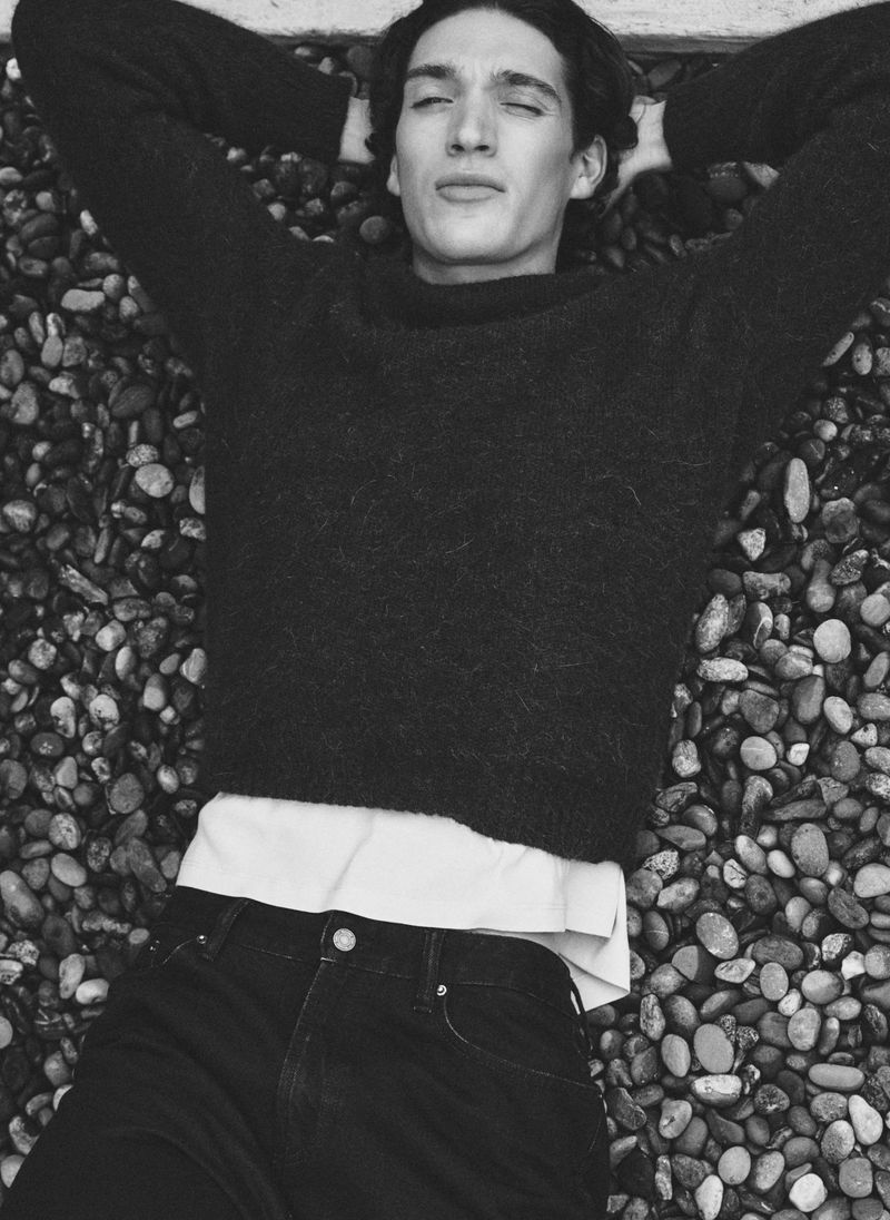Monochromatic portrait of a young adult man in a dark sweater and jeans lying on a bed of small rocks outdoors, with closed eyes.