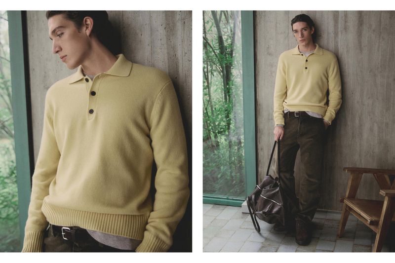 Indoor shot of a male model wearing a pale yellow polo sweater, brown trousers, and holding a duffel bag, leaning against a concrete wall.
