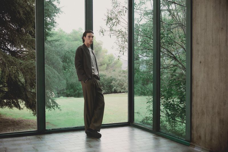 Young man in casual clothing poses in a modern indoor space with large windows overlooking a lush green landscape, fashion-forward aesthetic.