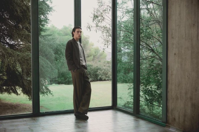 Young man in casual clothing poses in a modern indoor space with large windows overlooking a lush green landscape, fashion-forward aesthetic.