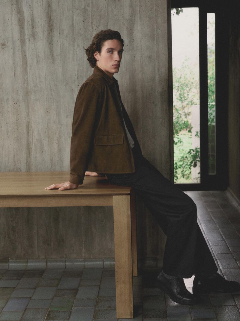 Young man seated on a wooden table wearing a brown jacket in a concrete interior for Massimo Dutti Field Notes campaign.