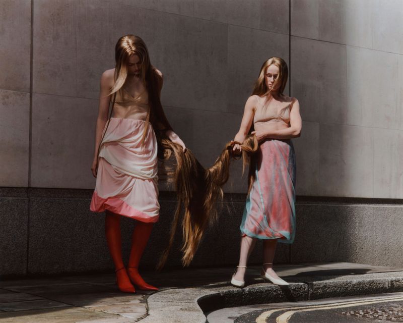 Two women with long hair stand in front of a grey concrete wall, wearing pastel-colored dresses and fashionable footwear on a city street.