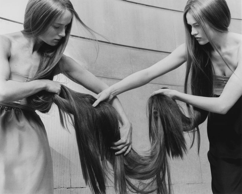 Monochrome shot features two light-skinned female models holding long, straight hair. One woman wears a light-colored dress; the other, a dark top.