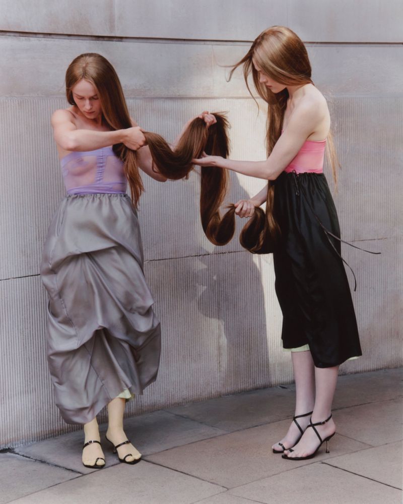Two light-skinned adult females wearing strappy heels hold long, flowing brown hair in front of a neutral wall, fashion editorial photography.