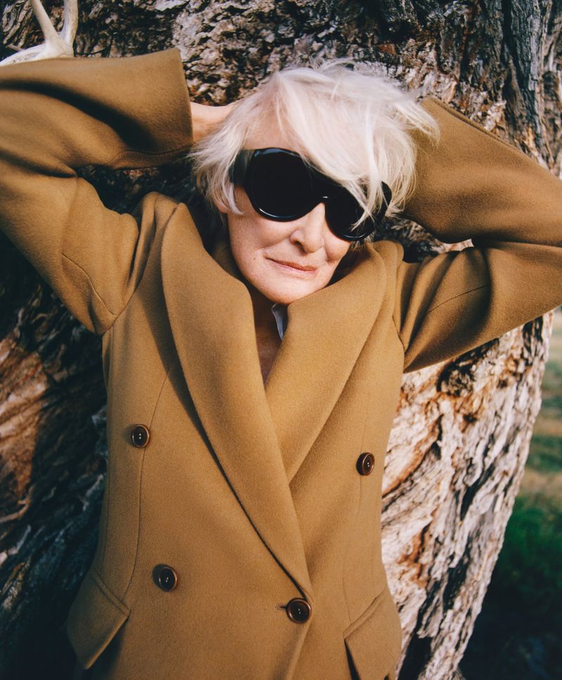 Glenn Close wearing oversized black sunglasses and a camel double-breasted blazer leaning against a large tree trunk.