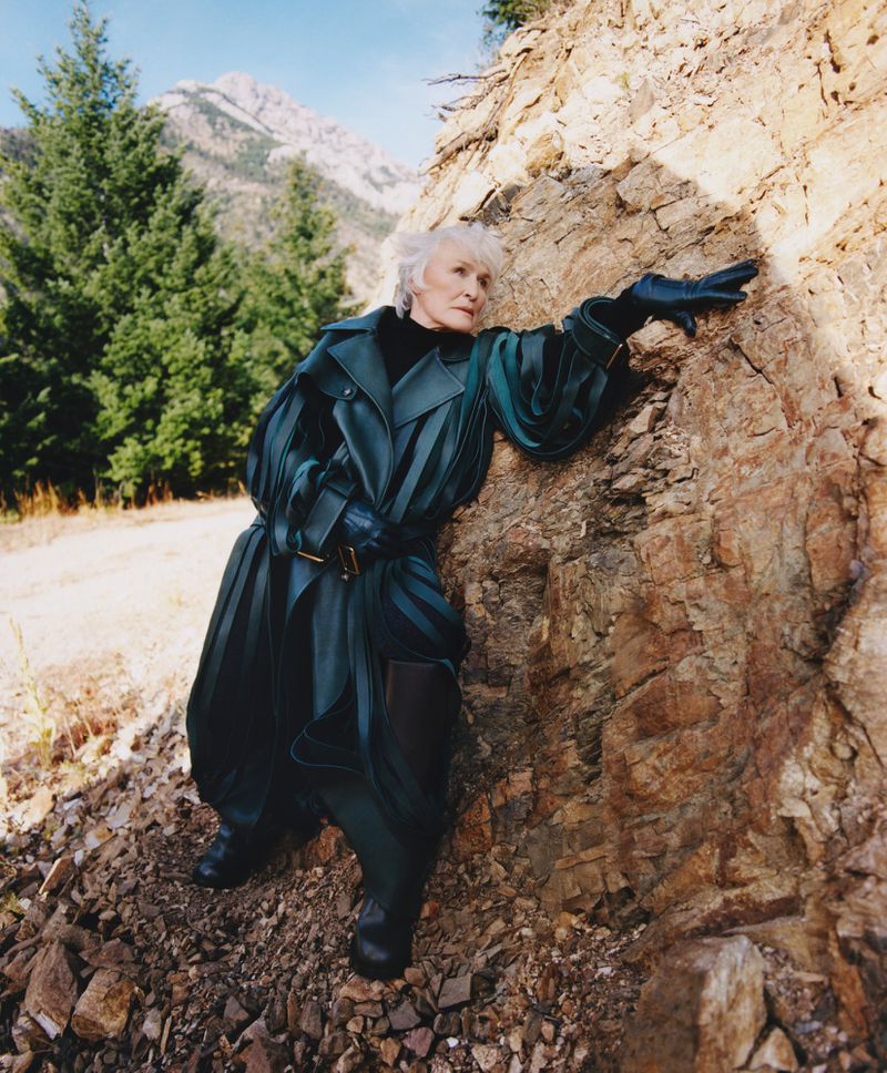 Elderly woman poses in teal trench coat near rocky terrain with mountains and trees in background, fashion-forward outdoor portrait.