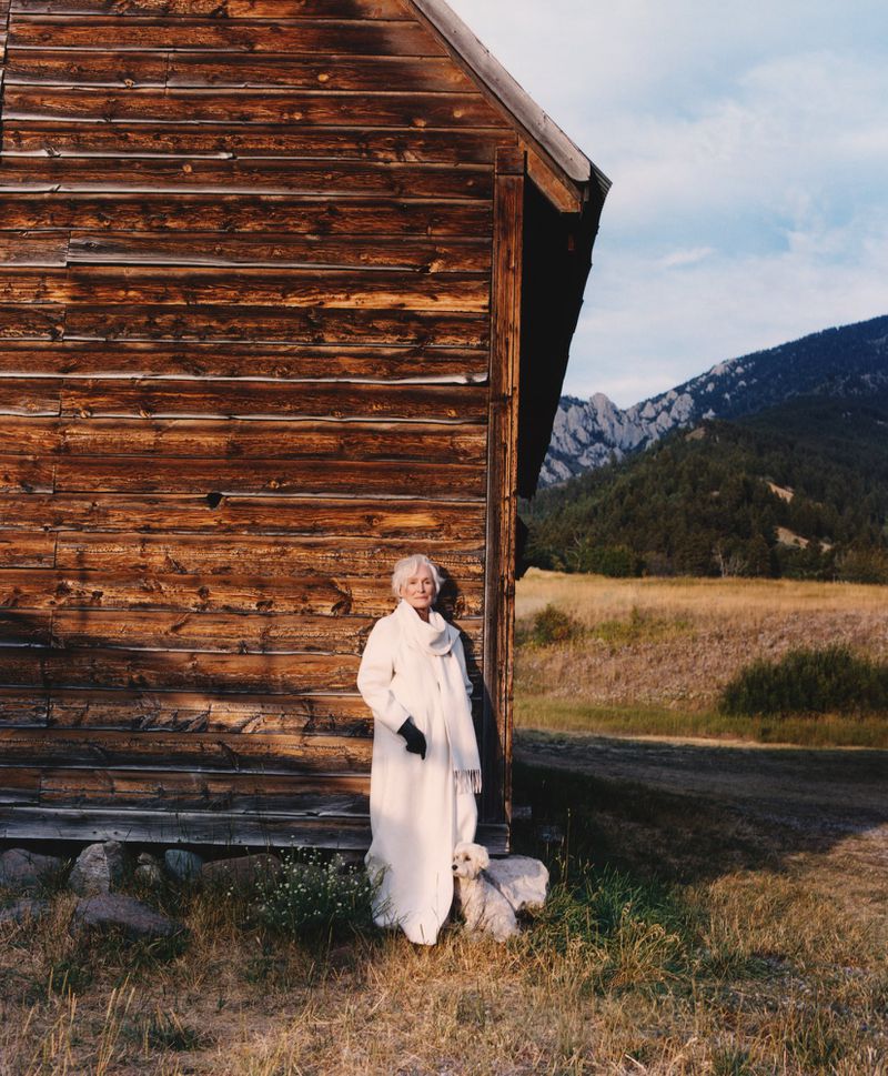 Elderly woman and a dog pose outdoors beside a rustic log cabin with mountains visible in the background under a partly cloudy sky.
