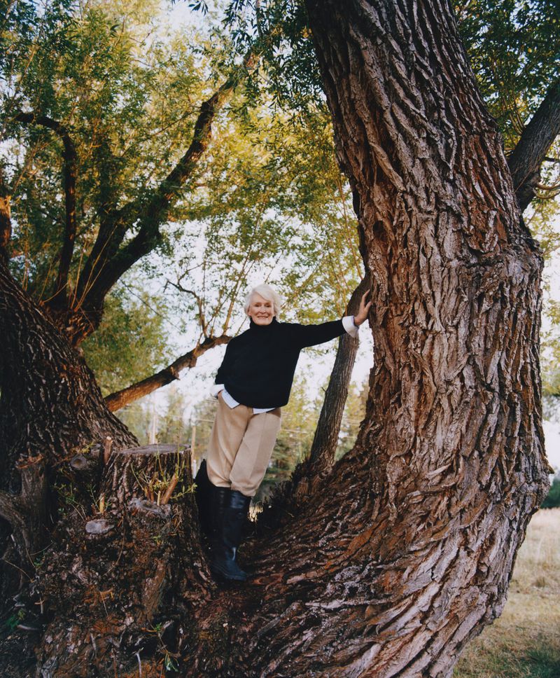 Woman with short white hair poses near a large tree with textured bark in an outdoor setting, wearing a black turtleneck and boots.