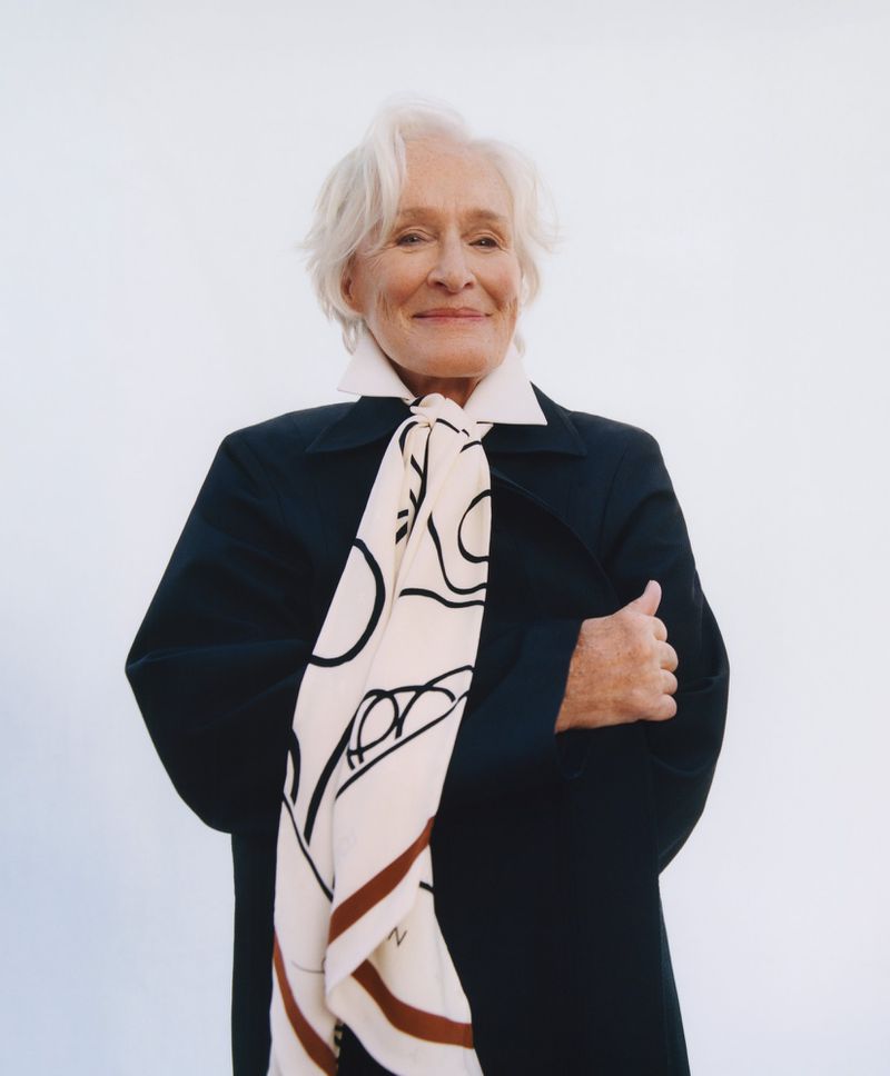 Actor Glenn Close smiling in a black coat and graphic patterned scarf against a white backdrop.