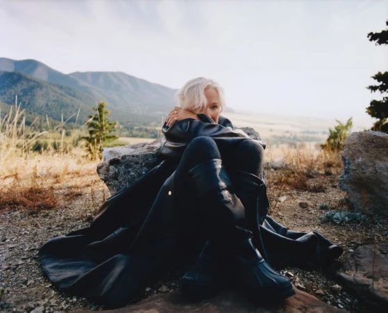 Mature woman sitting on rocks outdoors wearing a dark coat with mountains and sky in the background in an eye-level medium shot.