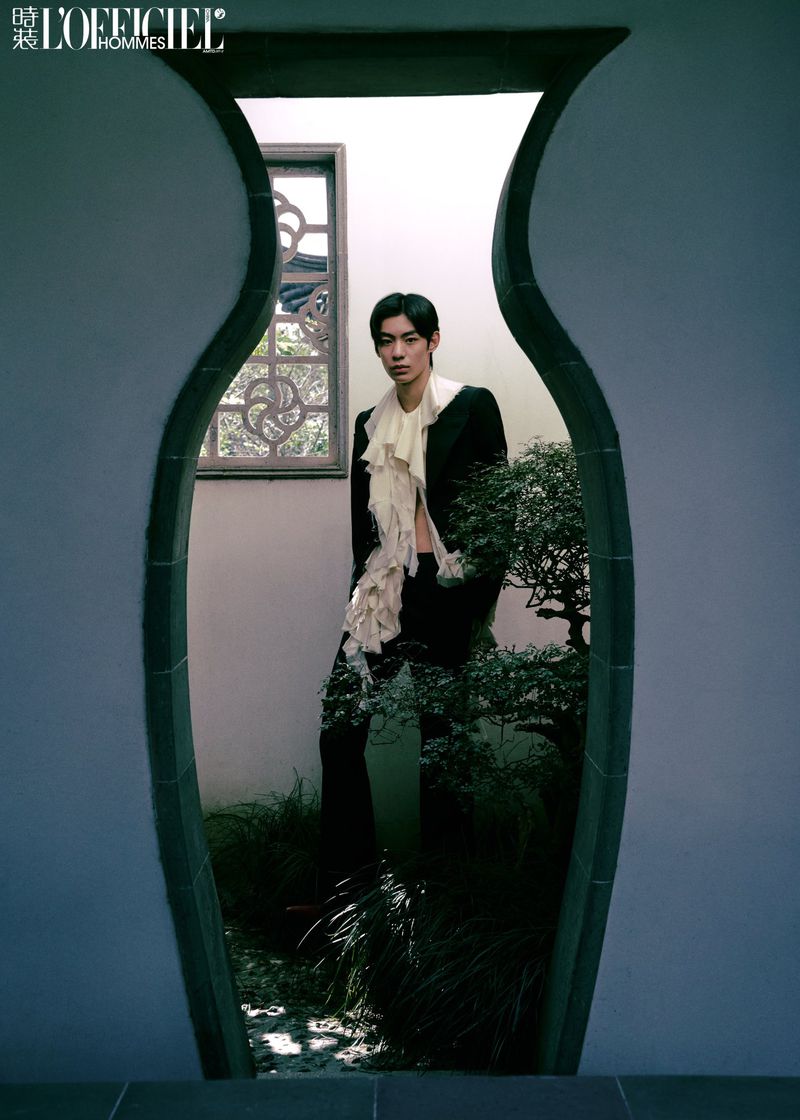 Asian male in a black suit with a ruffled white top framed by a decorative outdoor window surrounded by greenery and garden elements.